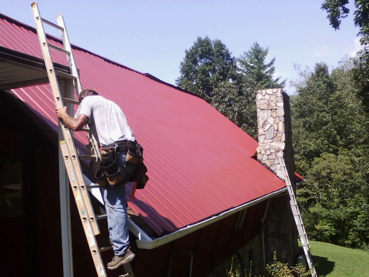 Roofer on a ladder beside a red metal roof and stone chimney — Soffit & Fascia Repair by Roofing Brothers in Ellettsville